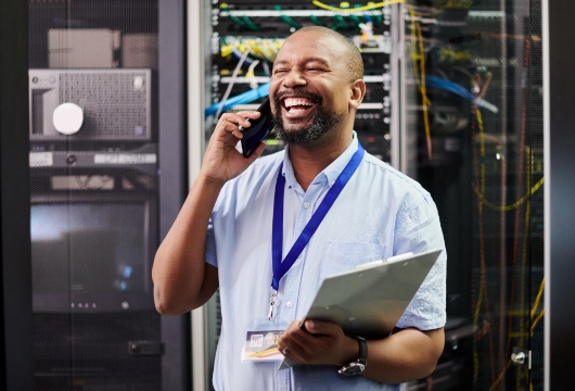 An image of a male engineer laughing on a cell phone in a data center holding a clipboard.