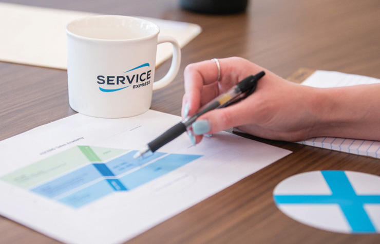 An image of a hand holding a pen to a paper with a chart on it. A Service Express mug is on the desk next to the paper.