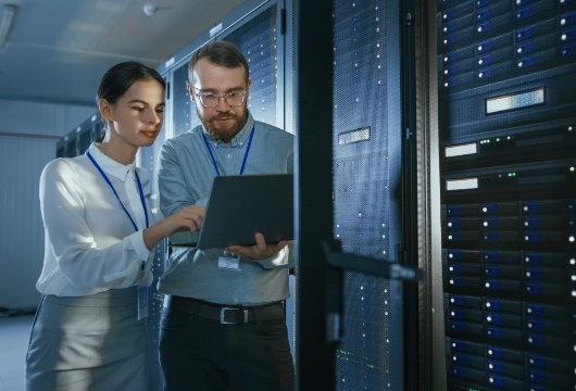 An image of a male and female engineer in front of an equipment rack in a data center. The male engineer is holding a laptop and the female engineer is pointing to the screen.
