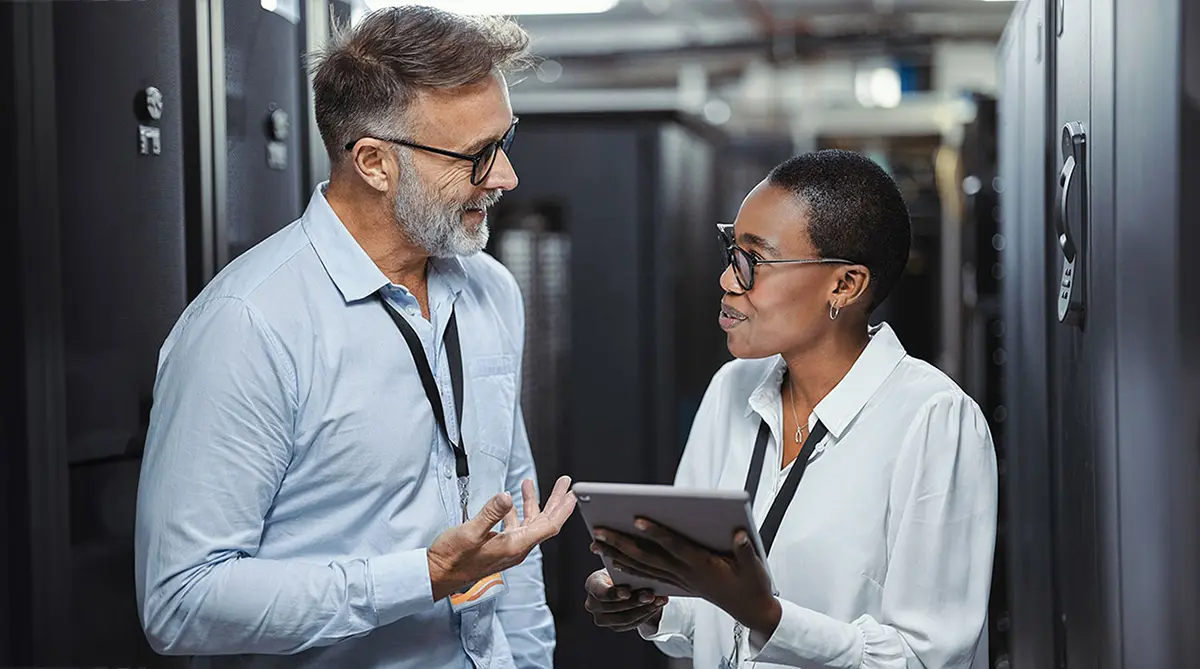 An image of two engineers in a data center. They are smiling and conversing. The female engineer is holding a tablet.