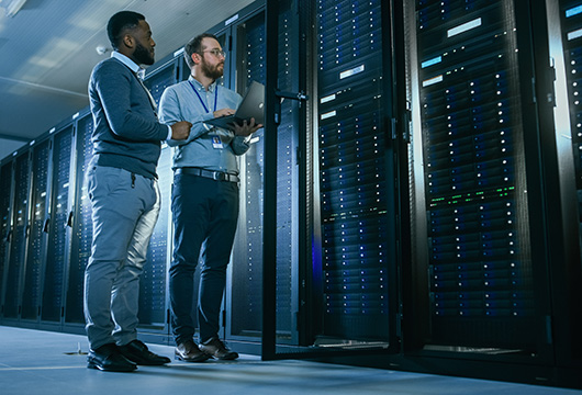 An image of two male engineers in a data center. One is holding a laptop and they are looking at equipment in a rack