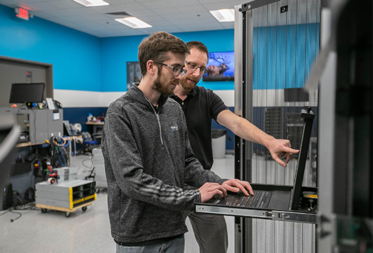 An image of two male engineers in a data center. One is pointing at a screen instructing the other engineer that is typing on the machine