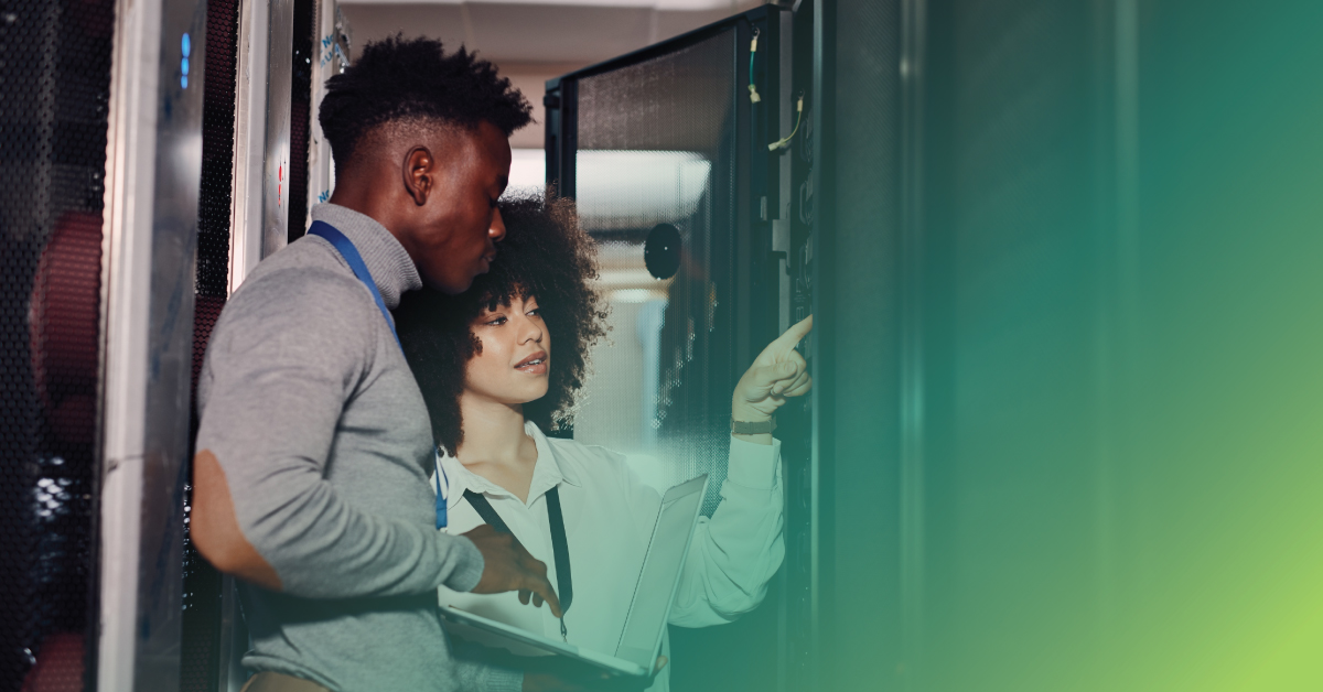 An image of a male and female engineer standing in front of a data center equipment rack. The female engineer is pointing to equipment within the rack.