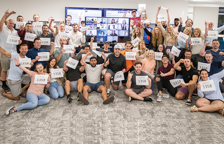 An image of a group of employees holding signs celebrating in front of a screen with employees on zoom