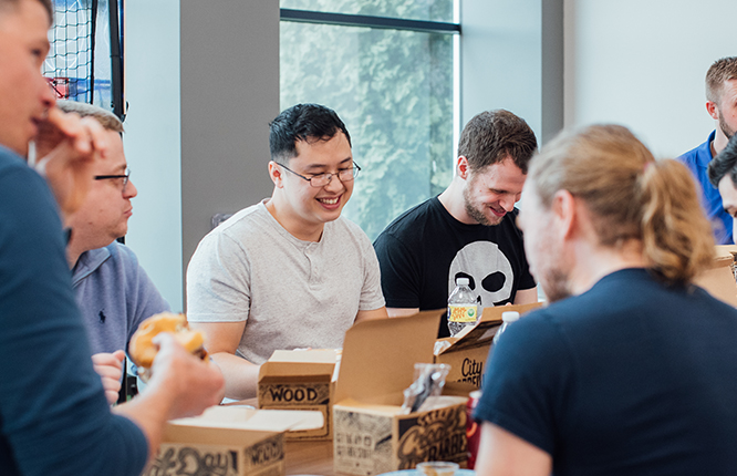 An image of coworkers gathered around a table with lunch boxes, eating, talking and smiling with each other