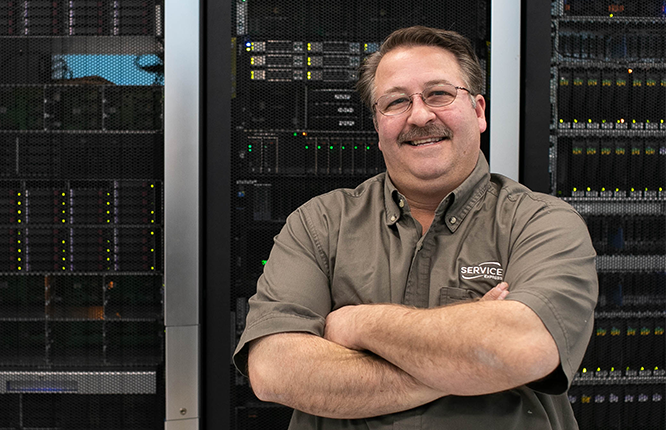 An image of an male engineer hands crossed around his chest smiling in front of a server