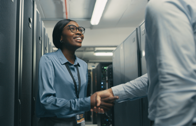 An Image of a female professional shaking the hand of someone just out of the shot on the right side. The two subjects are standing in a data center.