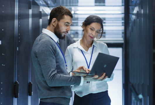 An image of a male and female in a data center. The male is holding a laptop with his hand perched on the trackpad. He is tilting the laptop toward the female so she can see the screen.