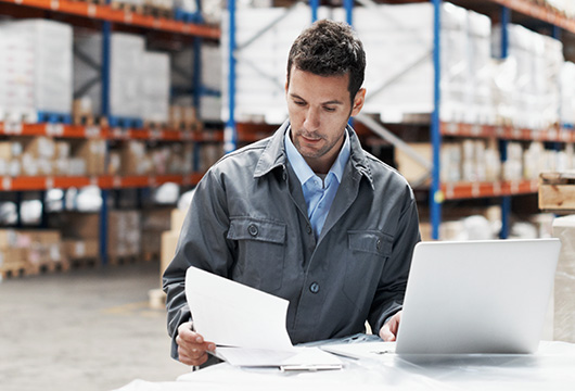 An image of a worker in a warehouse reviewing a paper and using a laptop