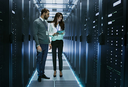 An image of a male and female engineer in a data center looking at equipment, the female engineer is holding a clipboard