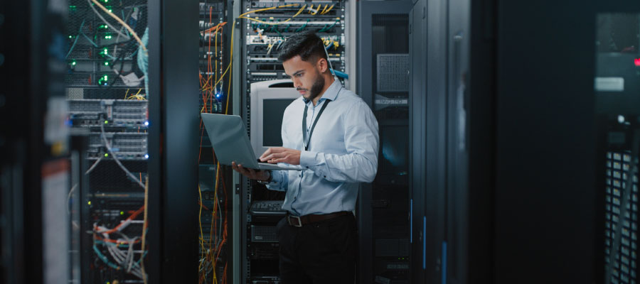 An image of a male engineer in a data center holding a laptop in one hand and typing with the other