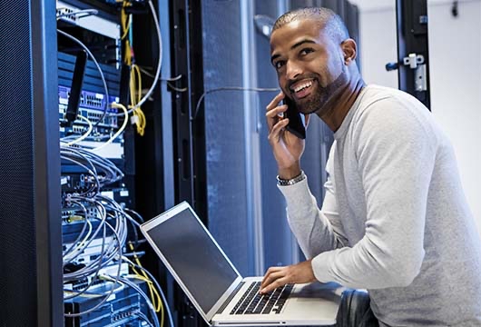 An image of an engineer in front of an equipment rack at a laptop holding a cell phone smiling